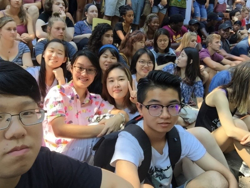 A group of international students sitting together in an assembly and smiling for the camera within a larger group.