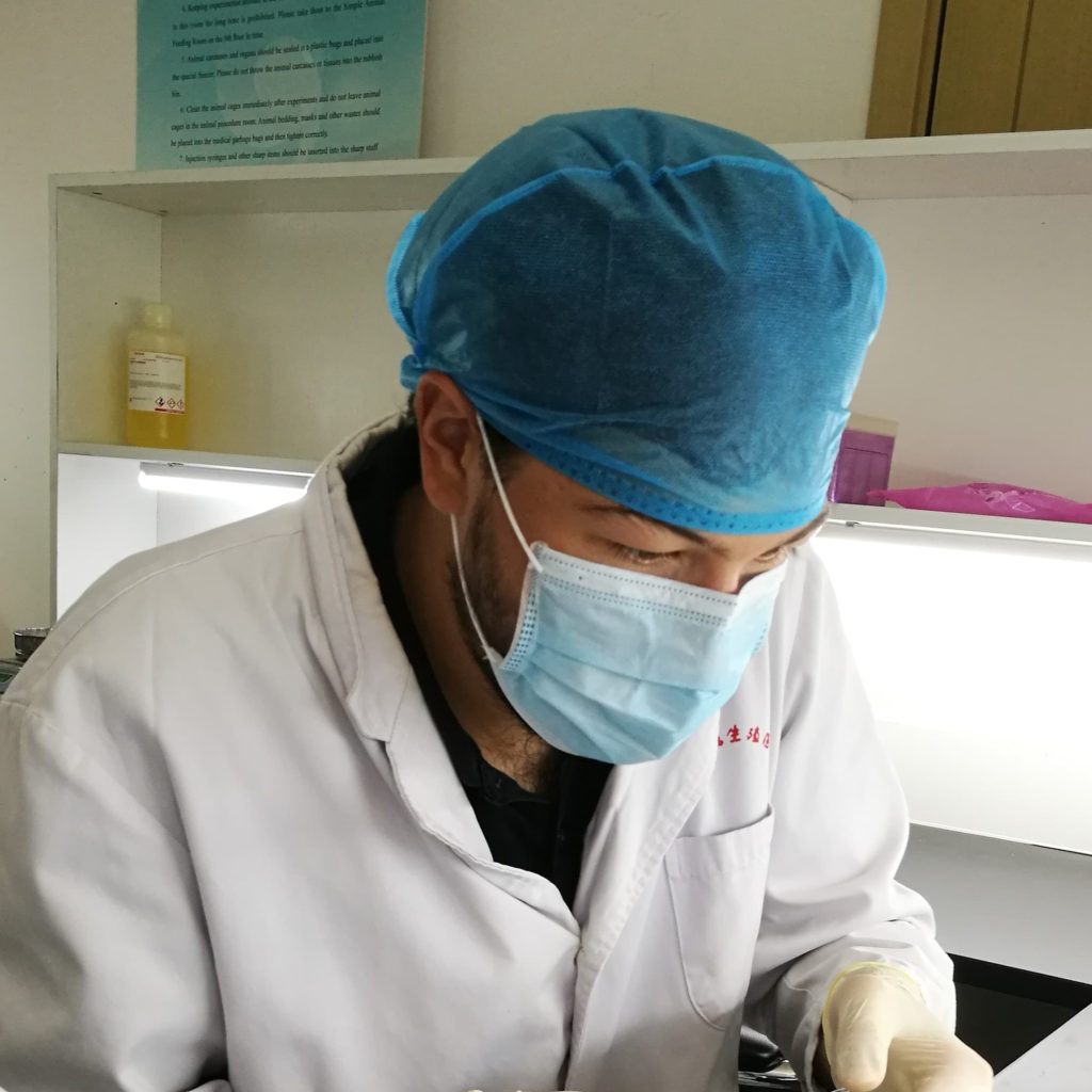 Man in lab wearing surgical head covering and face mask, earing a lab coat and gloves.  He is intently looking at his work.