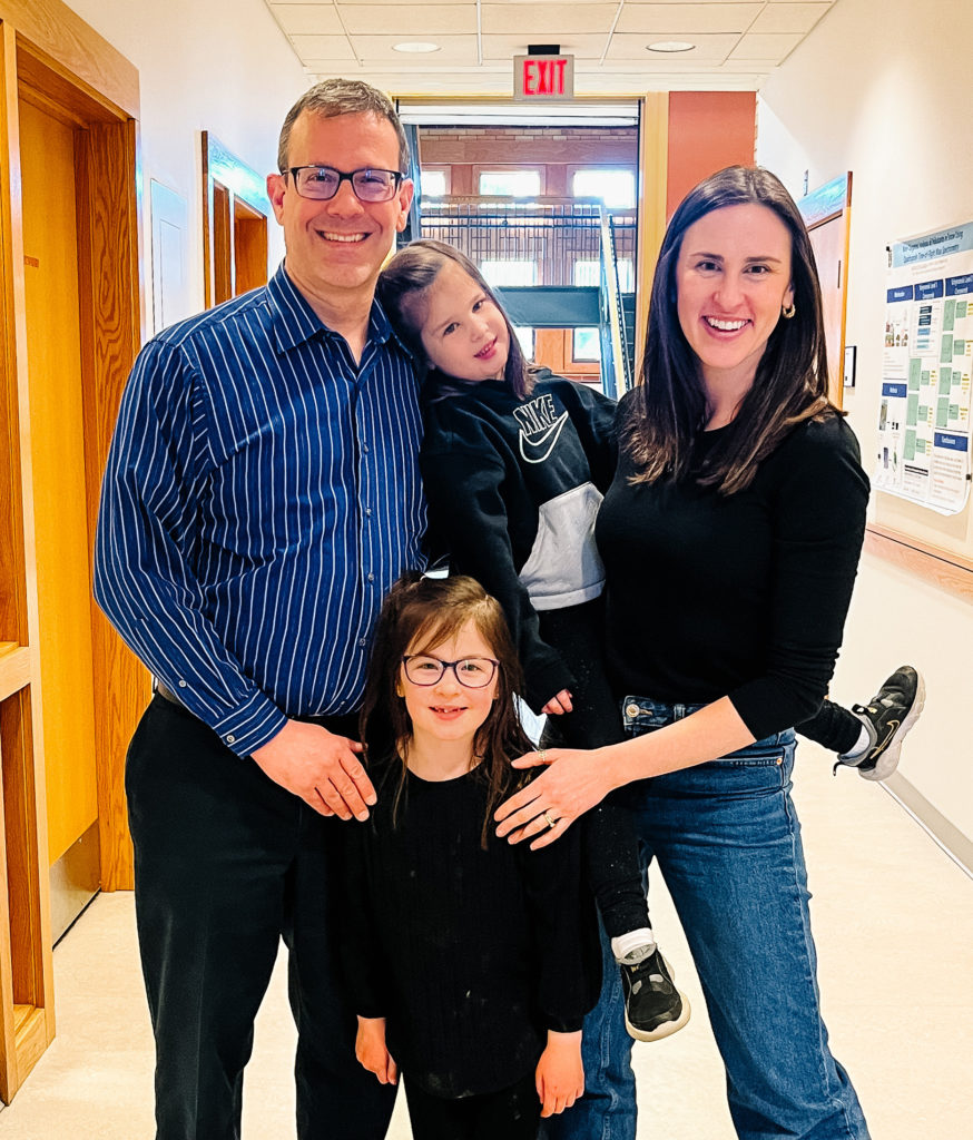 Professor standing next to adult holding a child. Standing in between the professor and adult is another child who is wearing glasses and smiling. Standing in the second floor hallway of Severance Hall.