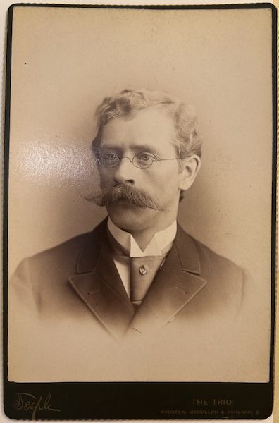 Sepia-toned cabinet-card portrait of William Z. Bennett, shown from the chest up, gazing slightly left. He wears pince-nez glasses, a full handlebar moustache, a high stand collar with cravat, and a dark double-breasted coat. A typed label along the right margin identifies him as a faculty member (“FACULTY — BENNETT, WILLIAM Z.”), and the studio imprint “THE TRIO, Wooster, Massillon & Ashland, O.” appears beneath the image.